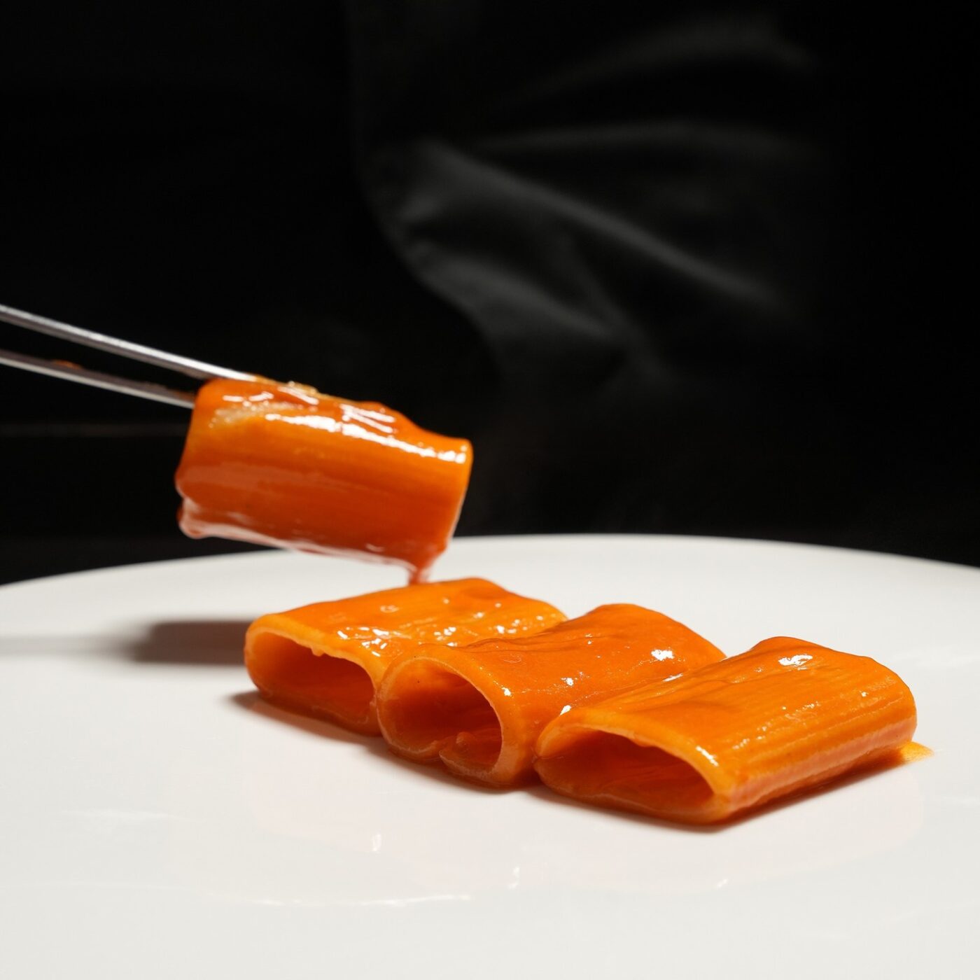 Paccheri allo scoglio - A close-up of four pieces of smooth, orange pasta arranged on a white plate against a dark background. A pair of chopsticks holds one piece above the plate. The pasta appears glossy and evenly colored.