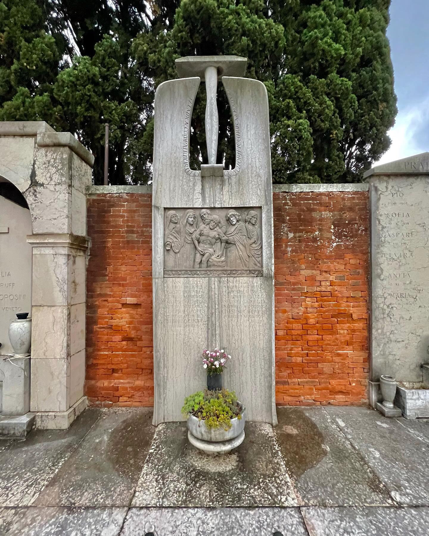 Un monumento in pietra con un intricato rilievo raffigurante figure umane appoggiate a un muro di mattoni. Sopra il rilievo c'è una scultura alta e astratta. Di fronte, c'è una piccola fioriera rotonda con piante verdi e fiorite. Alberi alti sono sullo sfondo.
