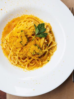 A plate of spaghetti topped with grated yellow bottarga and a sprig of parsley, placed on a brown table with a fork visible on the right side.