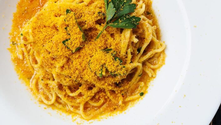 Certo! Con un titolo per l'immagine, il testo alternativo dovrebbe essere: Spaghetti alla Bottarga - Un piatto bianco di spaghetti con bottarga dorata grattugiata e guarnito con un rametto di prezzemolo, servito su un tavolo marrone con una forchetta parzialmente visibile sulla destra.