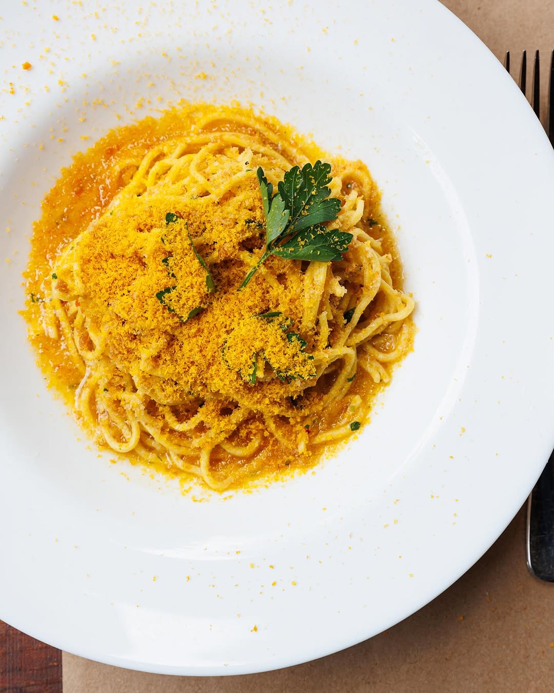 A plate of spaghetti topped with grated yellow bottarga and a sprig of parsley, placed on a brown table with a fork visible on the right side.