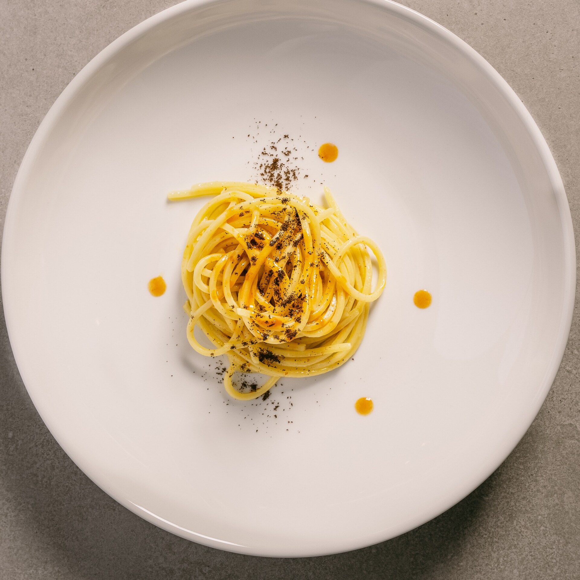 Sottovento Termoli - A minimalist presentation of spaghetti with a few strands placed in the center of a white plate, garnished with a sprinkling of black pepper and small droplets of orange sauce around it, on a gray surface.