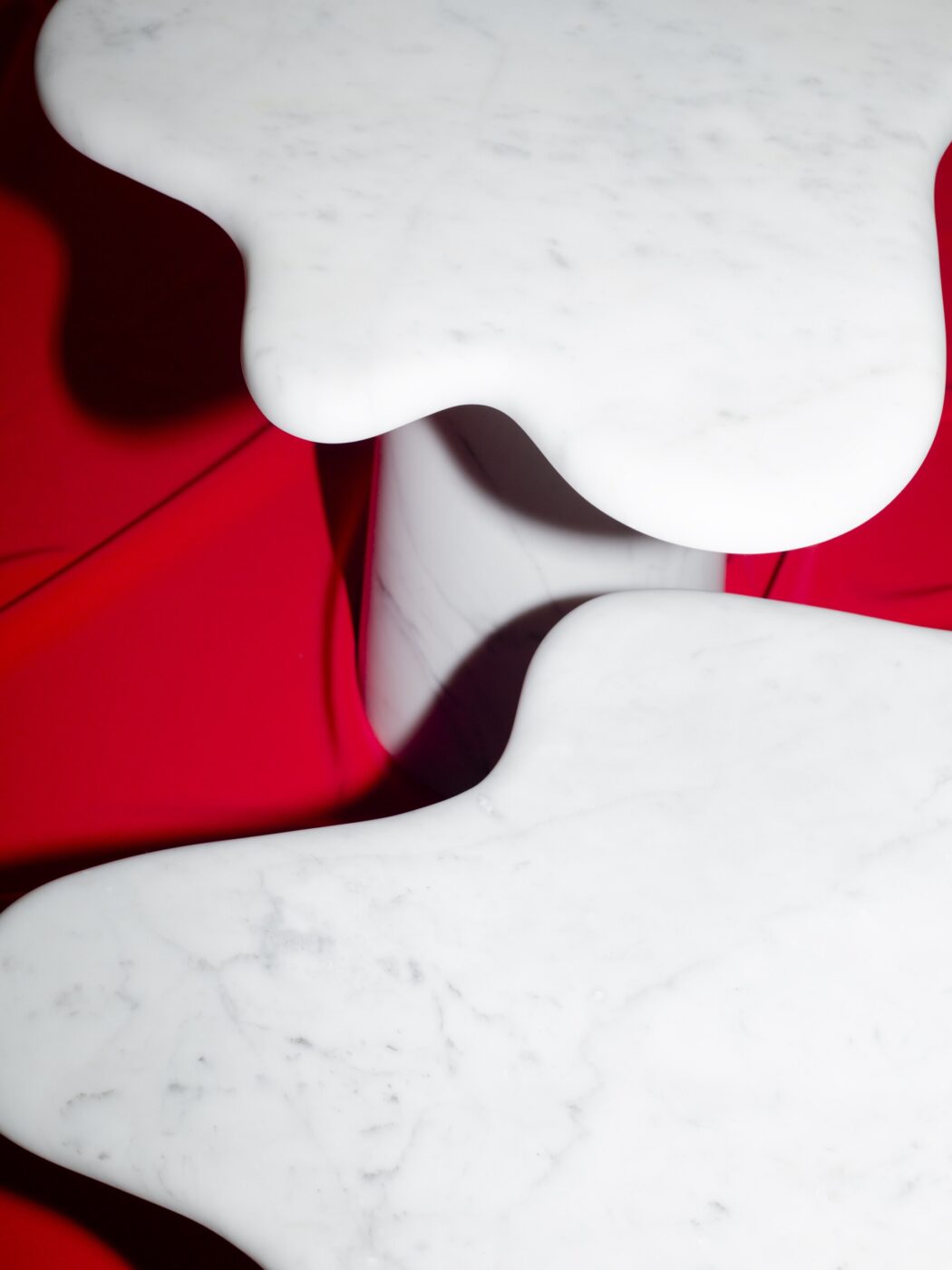 [Nessun titolo immagine] - Tavoli in marmo bianco con bordi irregolari e organici sono posizionati su uno sfondo rosso acceso, creando un contrasto sorprendente tra le superfici lisce e il colore intenso.
