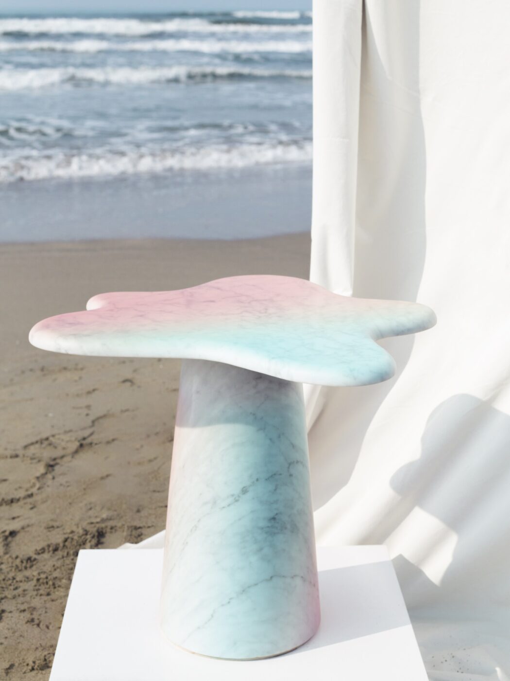 Un sogno pastello - Un tavolo in marmo color pastello dalla forma organica, simile a una nuvola, è posizionato su una spiaggia sabbiosa vicino all'oceano, con onde dolci e uno sfondo di tessuto bianco.