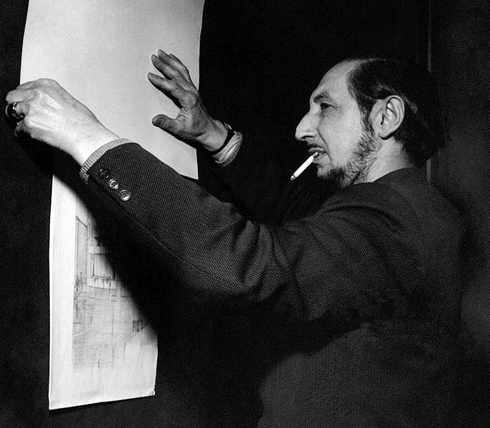 Titolo immagine - Testo alternativo: Una foto in bianco e nero di un uomo che tiene in mano un lungo foglio di carta con schizzi o disegni. Sta sistemando il foglio contro un muro e ha una sigaretta in bocca. Indossa una giacca da completo.