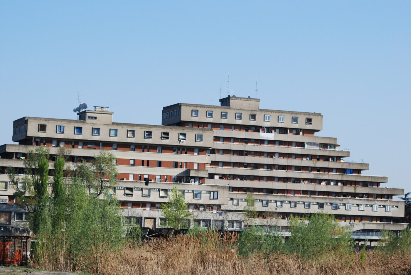 Brutalist Apartment Building - Condominium R-North, Brutalist-style apartment building with layered, staggered horizontal sections set against a blue sky. The structure features small windows and balconies. Trees and tall grass are visible in the foreground.