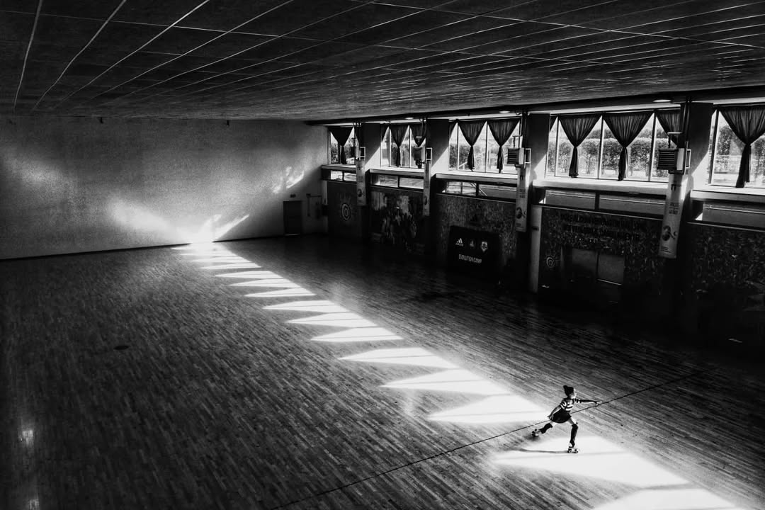 Corviale - Person roller skating in a large, empty indoor space with a wooden floor. Sunlight creates a zigzag pattern on the floor from windows on the right. The skater is positioned where the light and shadow meet, enhancing the dramatic effect.