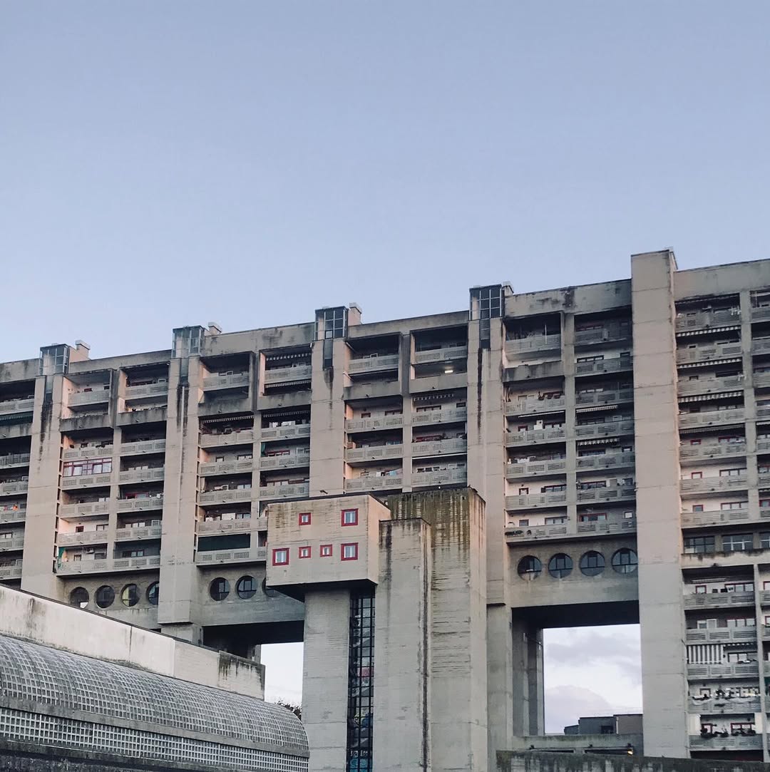 Rozzol Melara Complex - A large, brutalist-style concrete building stands against a clear blue sky. The structure features multiple floors with long horizontal windows and large, sturdy pillars at its base. Some greenery grows on the building.