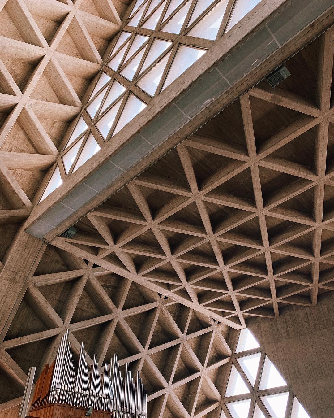 Santuario di Monte Grisa - Interior view of a modern church featuring a high, triangular-patterned wooden ceiling with large windows. Sunlight filters through, illuminating an organ in the corner. The architectural design showcases geometric shapes and natural materials.
