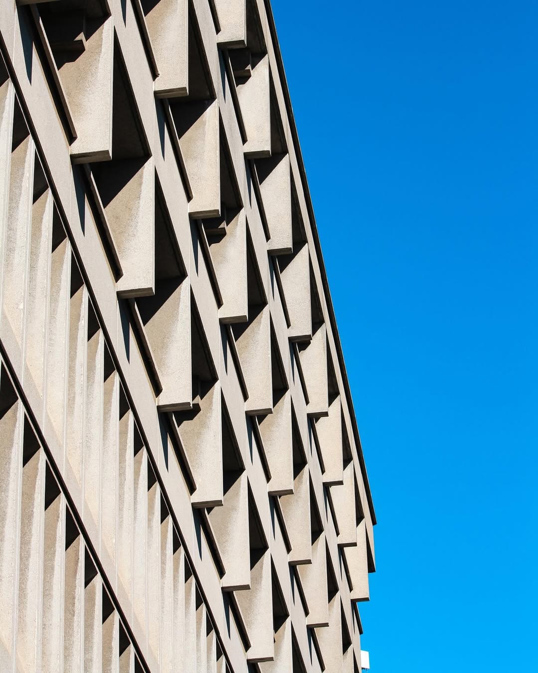 Bank of Italy Catania - Angled view of a beige concrete building with repetitive geometric patterns on its facade, set against a clear blue sky. The patterns create a series of triangular shadows across the building surface.