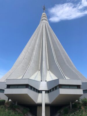 Basilica Santuario Madonna delle Lacrime - A modern, pointed concrete structure with a tall spire reaching into a clear blue sky. The building features geometric lines and windows near the base, creating a futuristic appearance. Sparse shrubbery surrounds the base.
