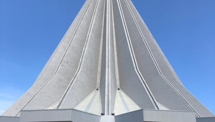 Futuristic Spire" - Un sorprendente edificio moderno con una struttura alta, bianca e conica sotto un cielo azzurro. Il design presenta linee eleganti e nette che conducono a una cima stretta, che ricorda una guglia futuristica. Una piccola struttura curva con finestre si trova alla base.