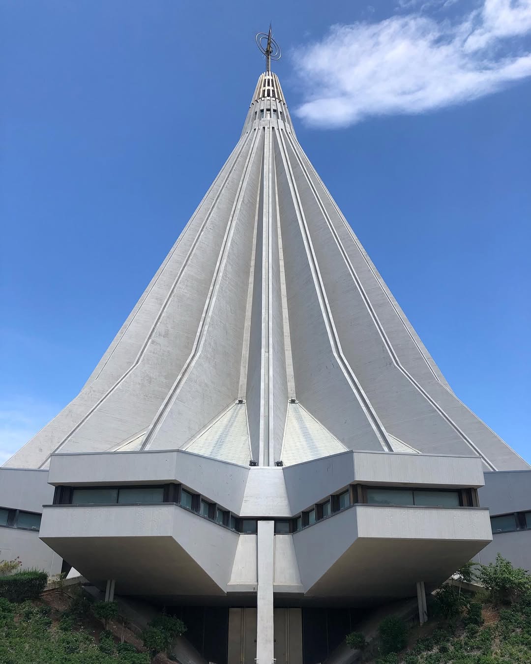 Basilica Santuario Madonna delle Lacrime - A modern, pointed concrete structure with a tall spire reaching into a clear blue sky. The building features geometric lines and windows near the base, creating a futuristic appearance. Sparse shrubbery surrounds the base.