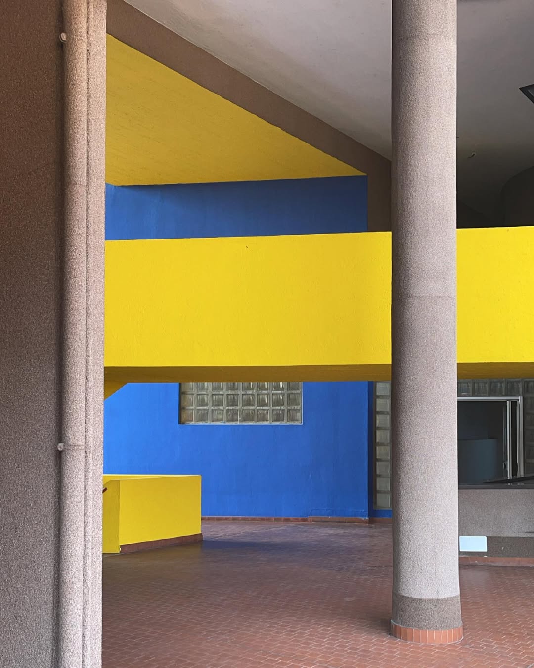 Monte Amiata - A modern architectural interior featuring a bold design with bright yellow walls and a deep blue accent wall. Tall cylindrical concrete pillars support the structure, and the floor is covered with reddish-brown tiles.