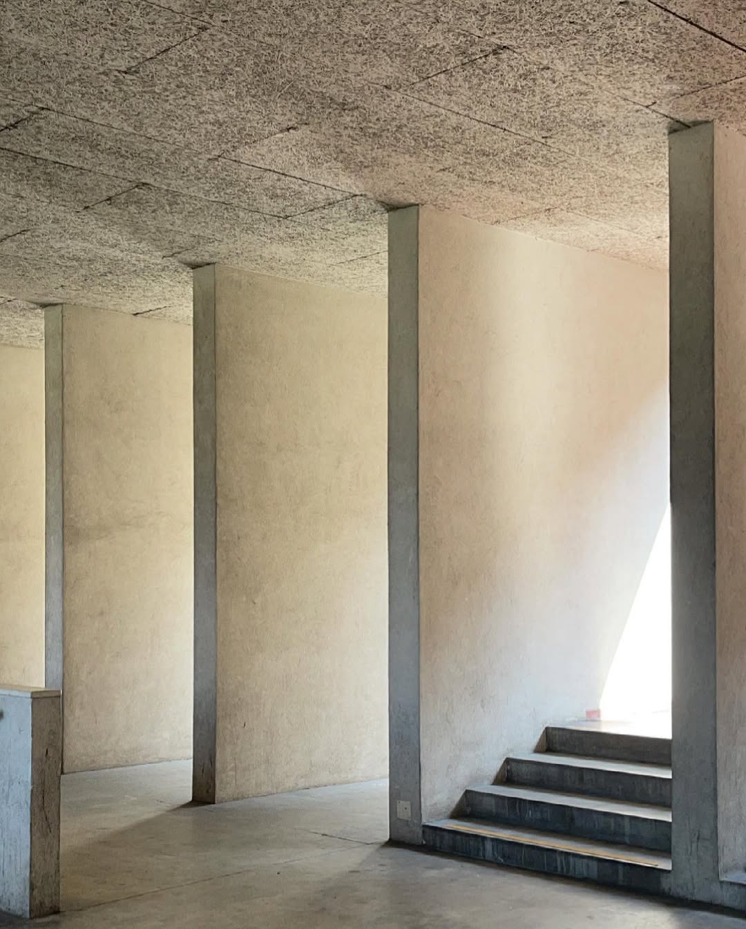 Monte Amiata - A minimalist interior features smooth, beige walls with several vertical beams. Sunlight streams through an unseen window, casting geometric light patterns on the walls and floor. Steps lead up to a well-lit area.