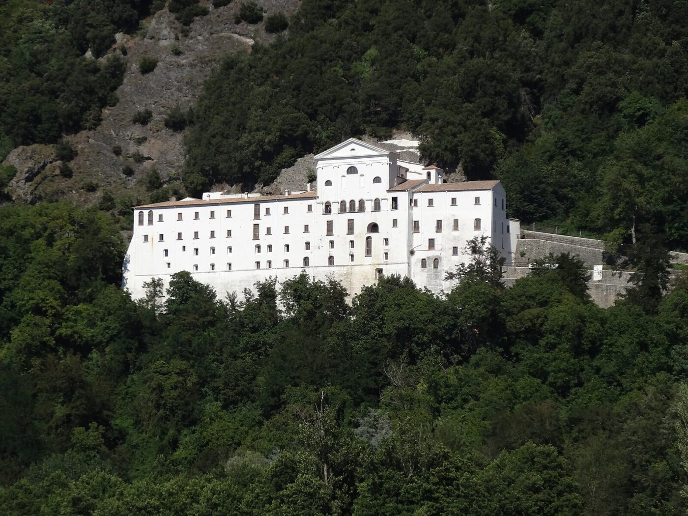 Un grande edificio bianco con numerose finestre, che ricorda un monastero o una struttura storica, sorge su una collina boscosa, circondato da rigogliosi alberi verdi e da un terreno roccioso.