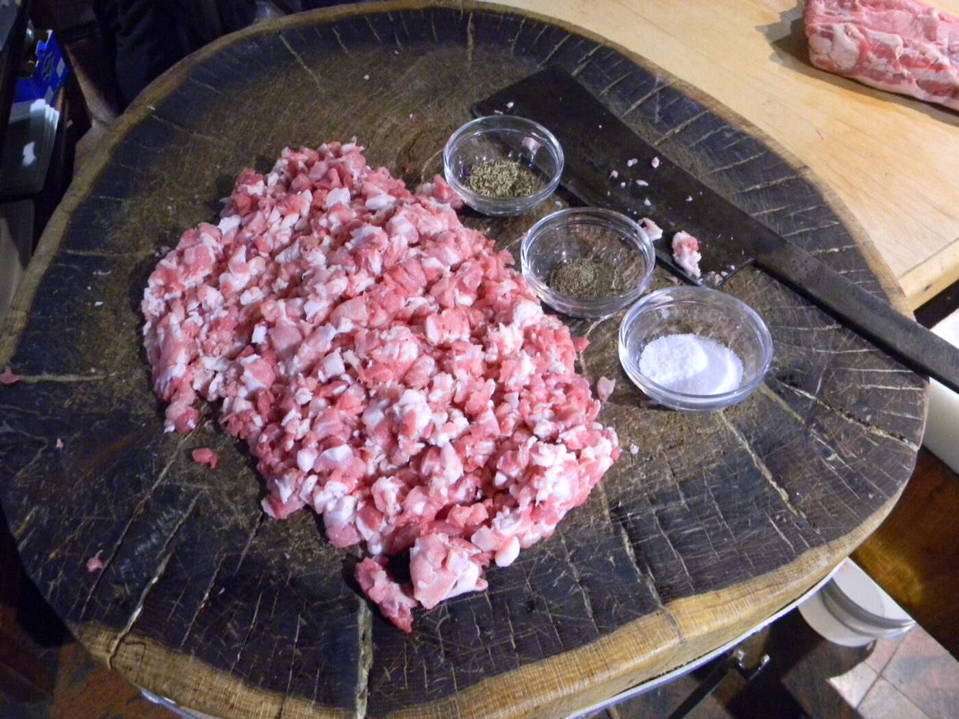 Chopped raw meat on a large wooden cutting board, with three small glass bowls containing pepper, herbs, and salt beside it. A knife rests near the bowls.