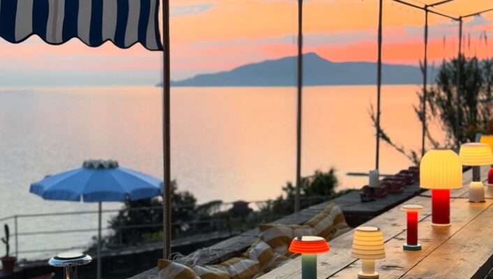 You haven’t provided an image title. If you want the alt text formatted as requested, please provide the image title. Assuming no image title is present, here’s your alt text: A wooden outdoor table with colorful lamps sits under a striped canopy, overlooking a calm sea at sunset. The sky glows with orange and pink hues, and there’s a blue umbrella and distant mountains in the background. If you’d like to add an image title for this scenario, let me know!.