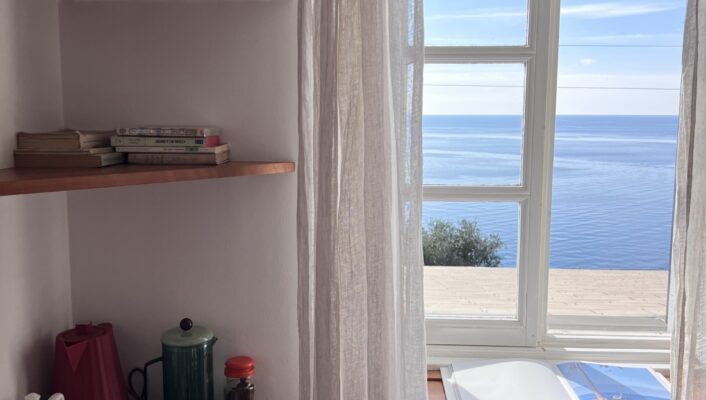 A cozy corner with wooden shelves holding books, a kettle, and cups beside a window with sheer curtains, overlooking a calm blue sea under a clear sky. An open book rests on the windowsill.