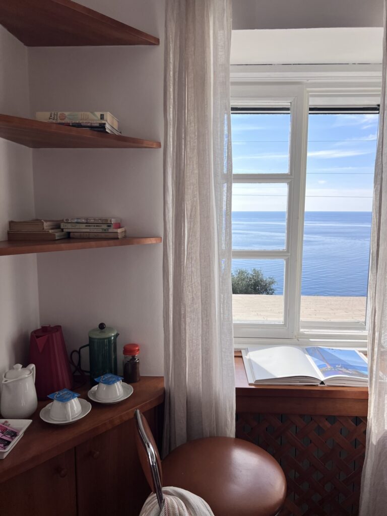 A cozy corner with wooden shelves holding books, a kettle, and cups beside a window with sheer curtains, overlooking a calm blue sea under a clear sky. An open book rests on the windowsill.