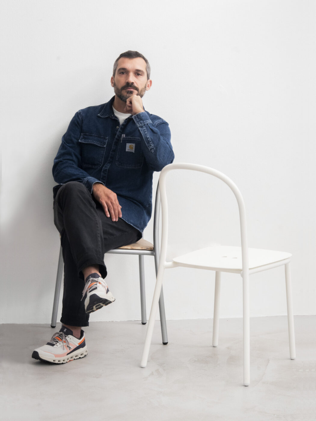 Certo! Ecco il testo alternativo formattato secondo le tue linee guida: [Titolo dell'immagine] - Un uomo con baffi e barba è seduto su una sedia contro una parete bianca, con il mento appoggiato sulla mano. Indossa una giacca di jeans, pantaloni neri e scarpe da ginnastica. Accanto a lui c'è una sedia bianca vuota.