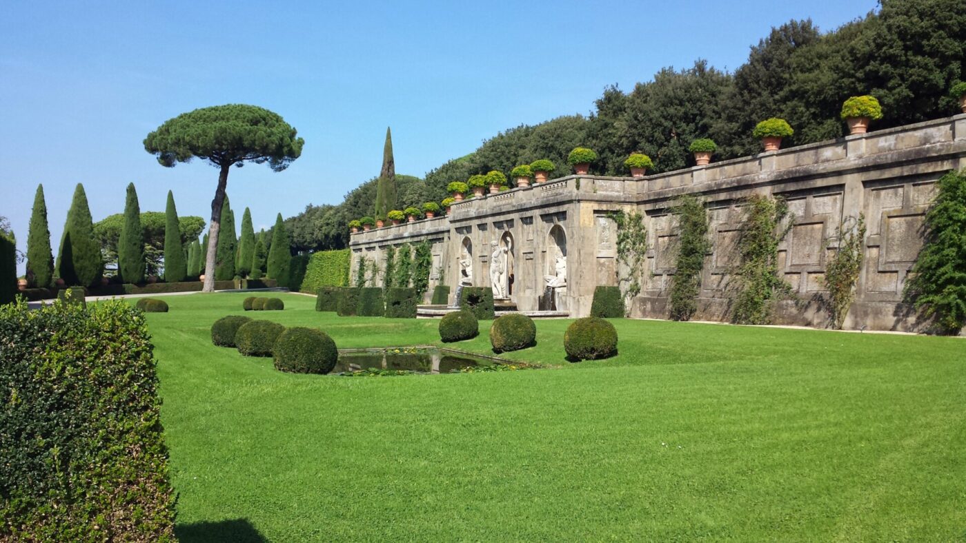 Un giardino formale con prati curati, siepi potate, arbusti rotondi, un piccolo laghetto e un muro di pietra ornato da statue e piante in vaso. Sullo sfondo si vedono alberi ad alto fusto e un cielo azzurro brillante.