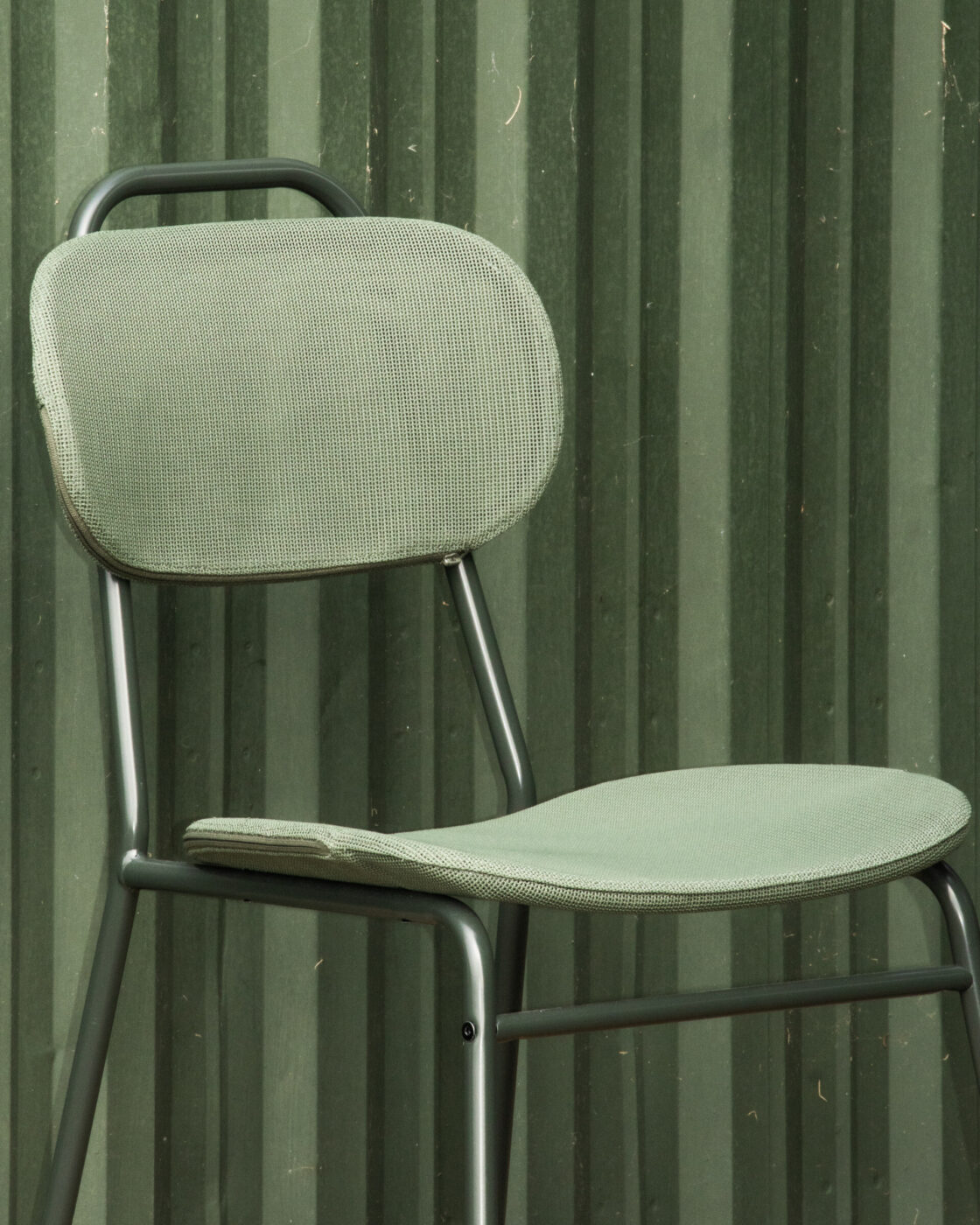 [Senza titolo] - Una sedia imbottita verde con struttura in metallo è posizionata di fronte a una parete a strisce verdi. La sedia e lo sfondo presentano tonalità simili, creando un effetto monocromatico.