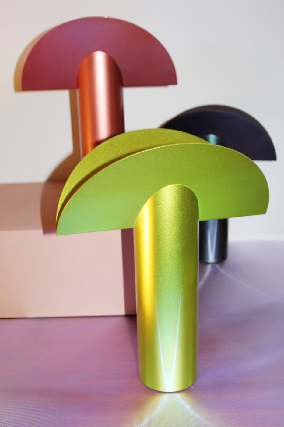 Tre moderne lampade metalliche a forma di fungo, nei colori verde, rame e nero, sono disposte su una superficie color lavanda e beige, con una luce soffusa che mette in risalto le loro texture lucide.