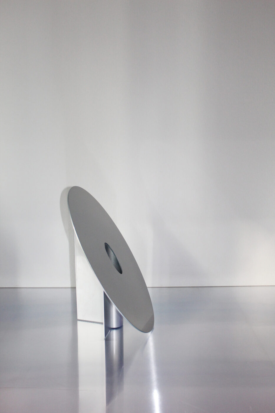 [Nessun titolo dell'immagine presente] - Una scultura metallica minimalista che presenta un grande disco inclinato con un foro centrale, sostenuto da una breve base cilindrica, su uno sfondo bianco.