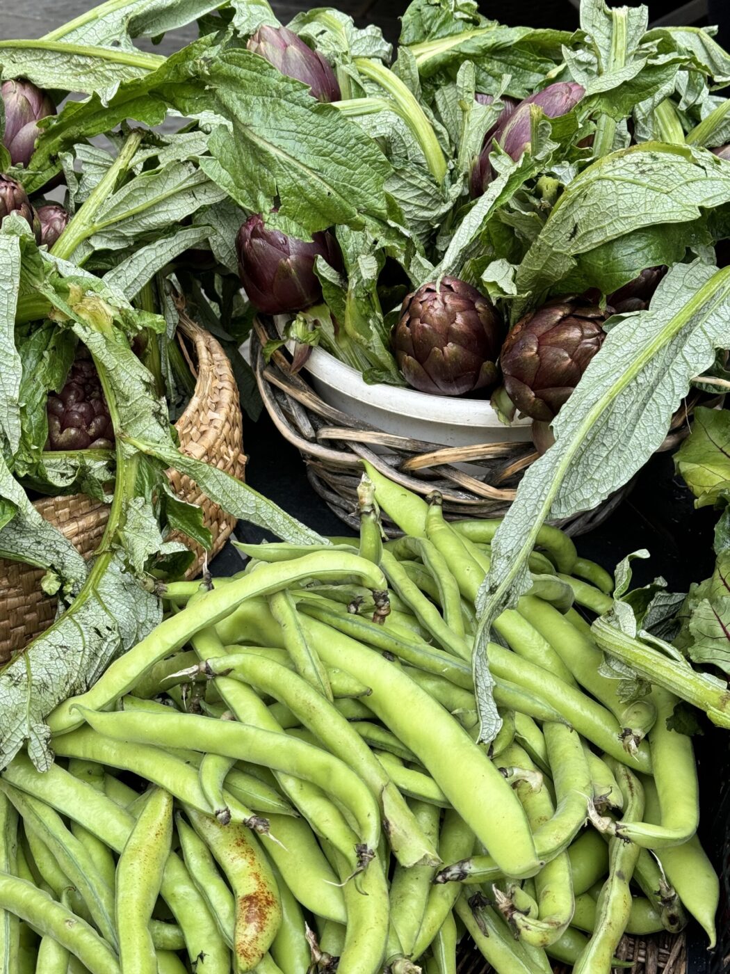 Baskets of Artichokes and Fava Beans - Baskets filled with fresh purple artichokes with leafy stems, and a pile of green fava beans displayed at a market.