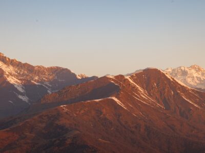 Mountain Range at Sunset - Mountain range under a clear sky at sunset, with warm sunlight casting an orange glow on the peaks and some snow visible on the higher elevations.