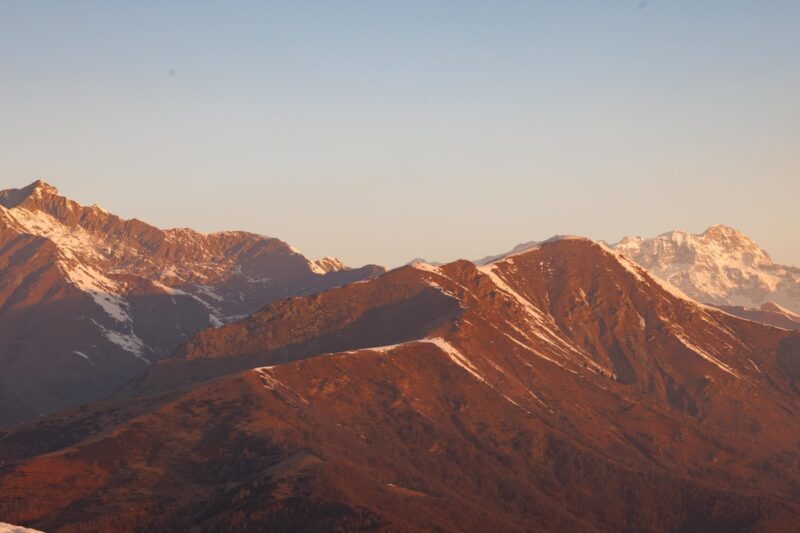 Mountain Range at Sunset - Mountain range under a clear sky at sunset, with warm sunlight casting an orange glow on the peaks and some snow visible on the higher elevations.