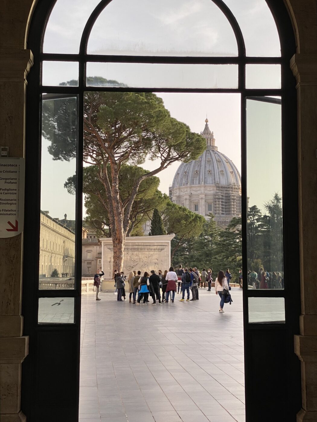 Non è stato fornito alcun titolo per l'immagine. Ecco un testo alternativo adatto: Un gruppo di persone è in piedi in un cortile incorniciato da una grande finestra ad arco, con un alto albero verde e la cupola della Basilica di San Pietro visibili sullo sfondo. La scena è immersa in una soffusa luce diurna.