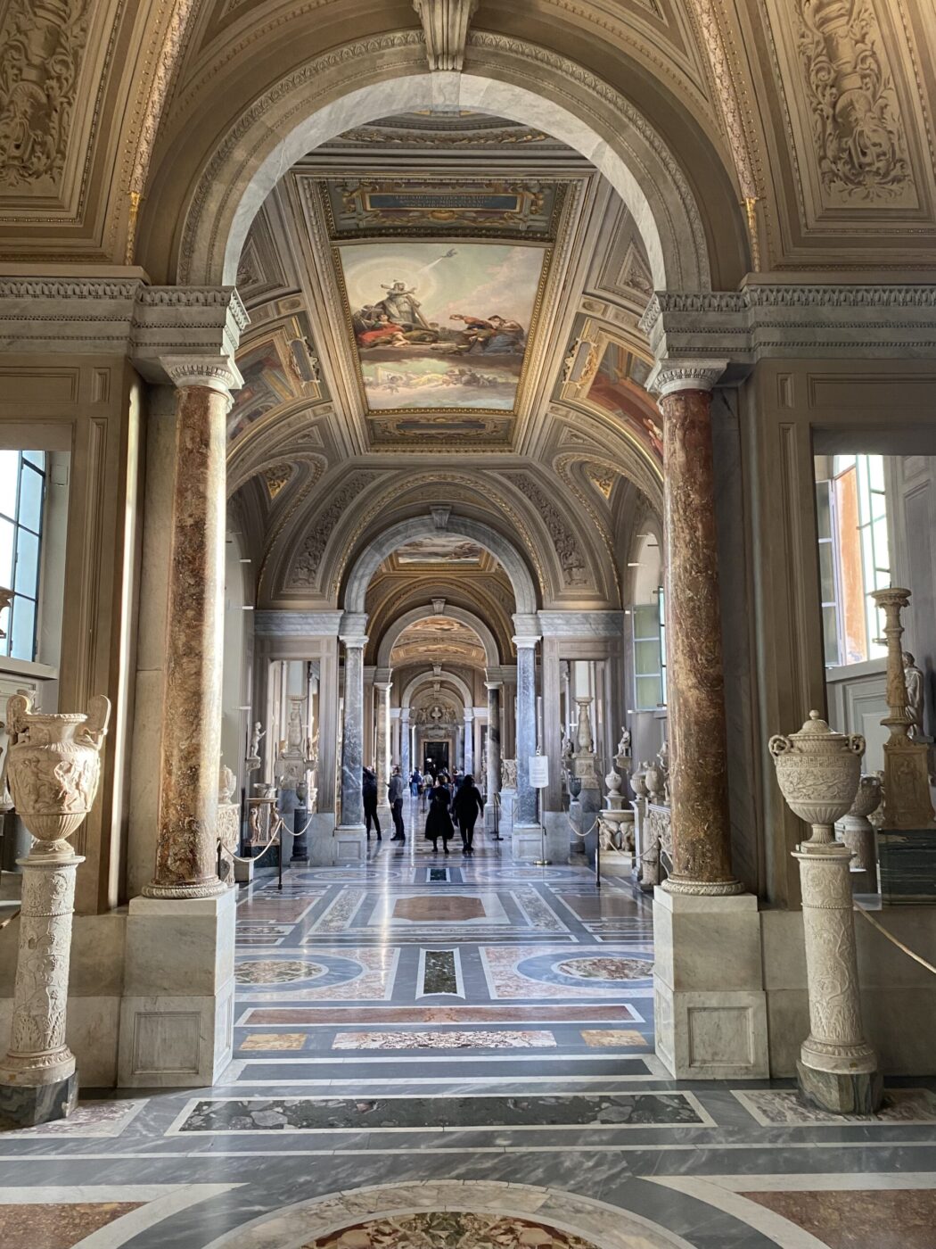 Un grande corridoio con soffitti a volta, colonne ornate, pavimenti in marmo, sculture classiche e intricati dipinti sul soffitto, con persone che camminano in lontananza sotto una soffusa luce naturale.