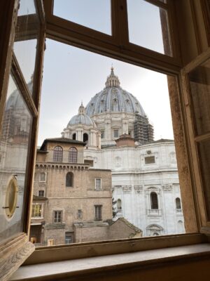 St. Peter’s Basilica View - View through an open window showing the dome of St. Peter’s Basilica, with nearby historic buildings and part of the basilica’s facade visible against a clear sky.