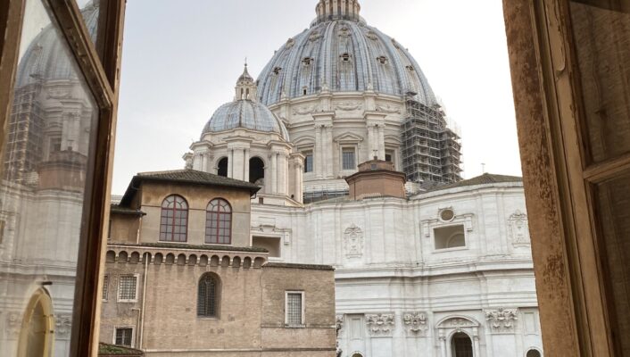 St. Peter’s Basilica View - View through an open window showing the dome of St. Peter’s Basilica, with nearby historic buildings and part of the basilica’s facade visible against a clear sky.