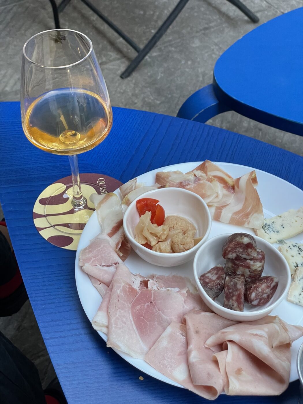 [Titolo dell'immagine] - Un bicchiere di vino color ambra è su un tavolo blu accanto a un piatto di salumi assortiti, tra cui fette di prosciutto, salame e formaggio. Un piccolo piatto con un contorno di peperone rosso o sottaceti è anche sul piatto.