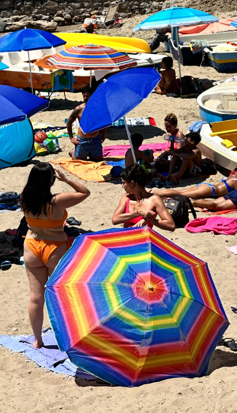 Liguria Beach : People relax on a sandy beach under colorful umbrellas. - Alt Text: In the foreground, a rainbow-striped umbrella stands beside two women in swimsuits. Other people are sunbathing or sitting under blue and orange umbrellas in the background on a sandy beach.