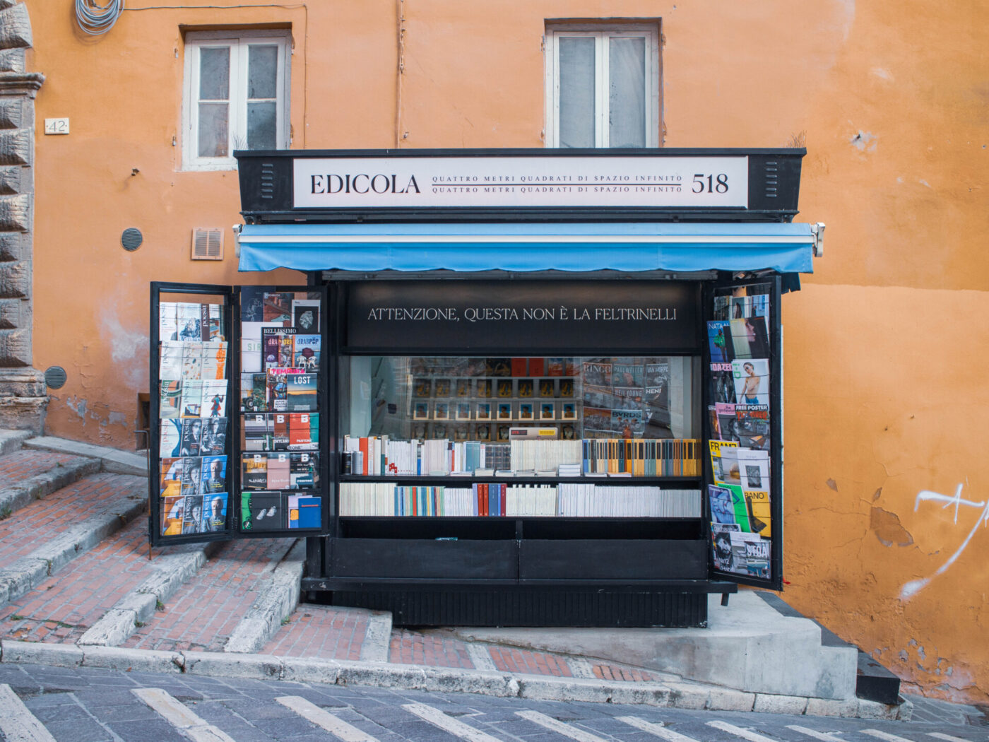 EDICOLA 518" - Un piccolo edificio arancione all'angolo ospita un chiosco con una tenda da sole blu con la scritta "EDICOLA 518". È pieno di libri e riviste e ha un cartello che dice "ATTENZIONE, QUESTA NON È LA FELTRINELLI". Gradini in mattoni conducono all'ingresso.