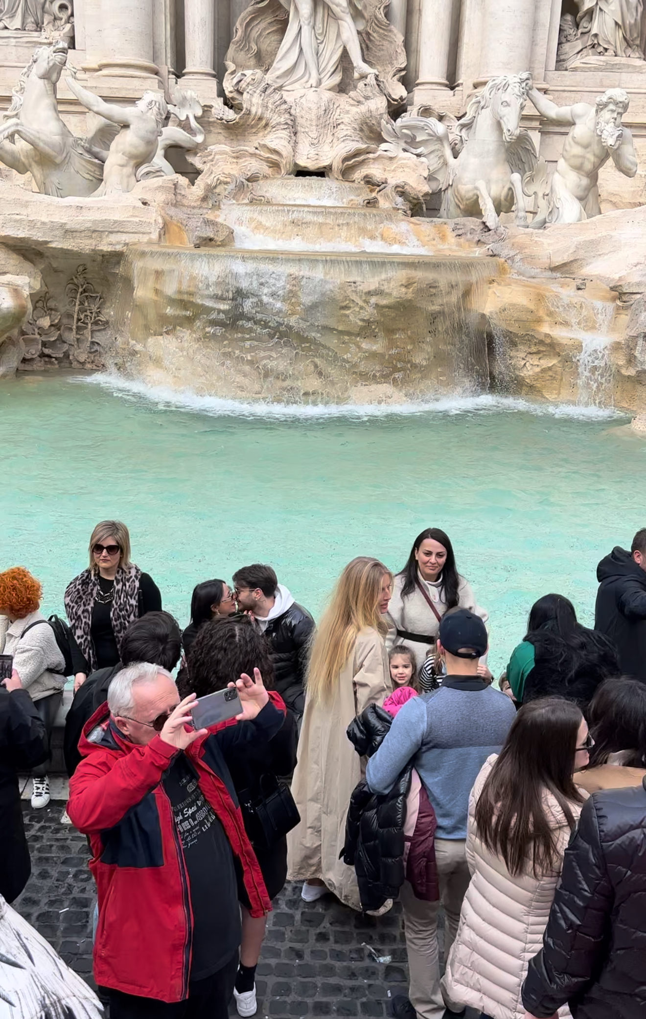 Trevi Fountain, Rome - A crowd of people gather in front of the Trevi Fountain in Rome, some posing for photos and others taking pictures. The turquoise water and sculpted stone figures of the fountain are visible in the background.
