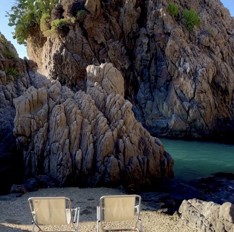 Due sedie a sdraio vuote di fronte a una caletta turchese - Due sedie a sdraio vuote sono adagiate su un terreno sabbioso, di fronte a una piccola caletta con acqua turchese e grandi formazioni rocciose frastagliate sotto un cielo azzurro e limpido. Rare piante verdi crescono sulle rocce.