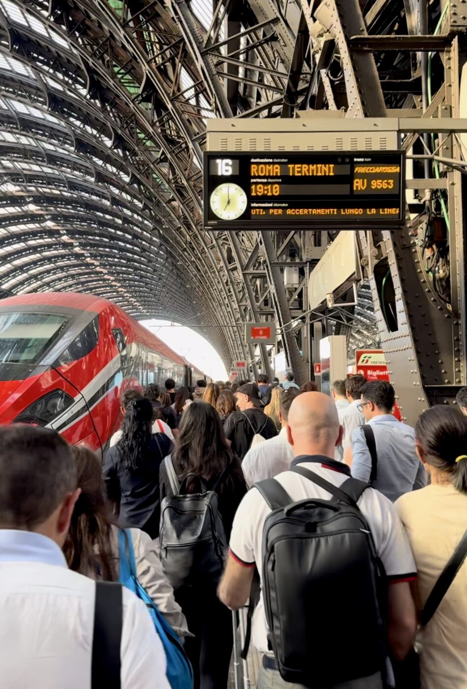 Roma Termini - A crowd of people walks toward a red and white high-speed train at an arched, covered station. An overhead sign displays Roma Termini and the departure time, with steel beams and lights framing the busy scene.