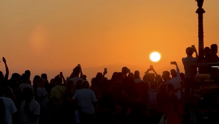 A crowd of people watches and takes photos of the sunset, silhouetted against an orange sky. A vintage street lamp stands on the right side of the image.