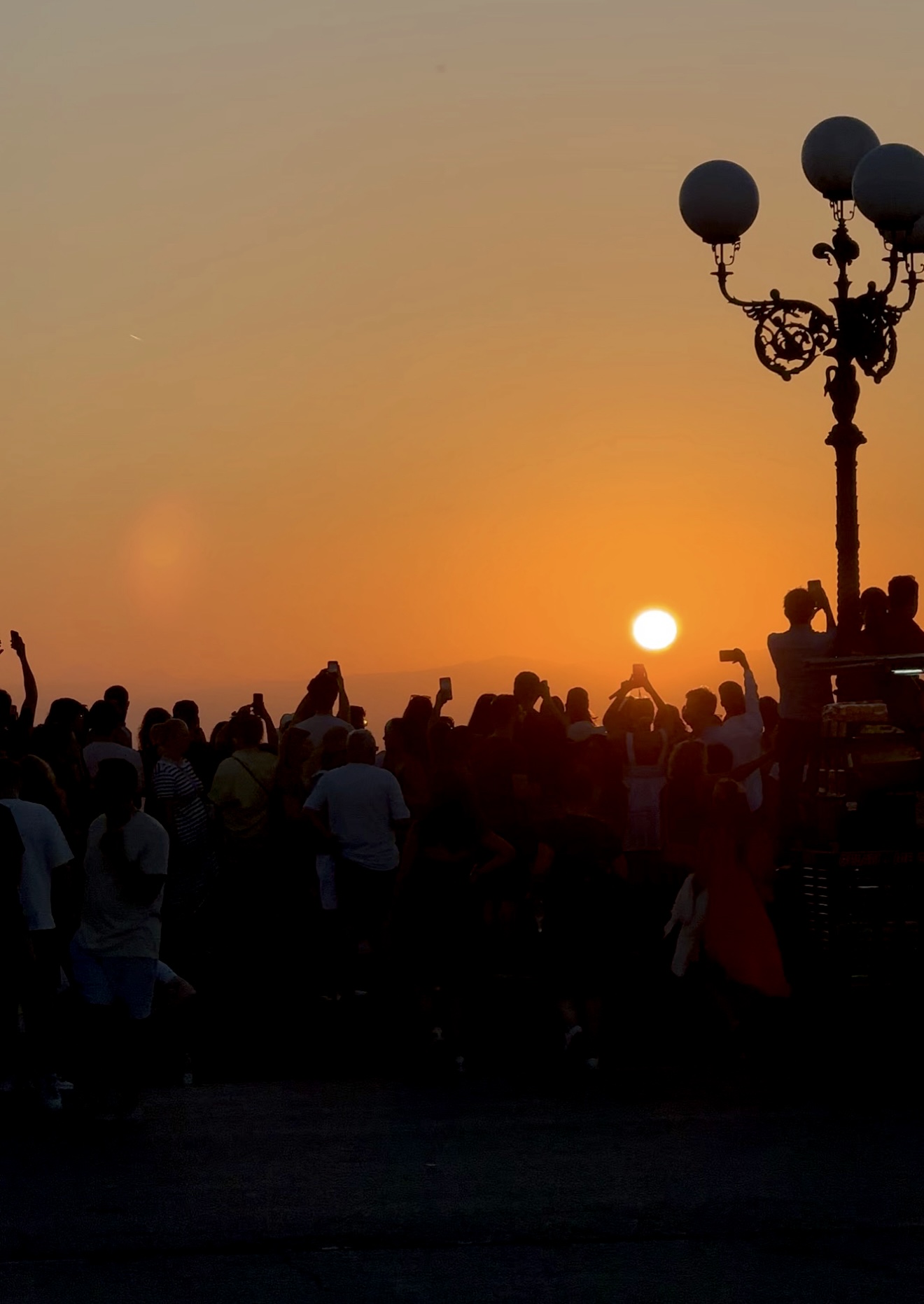 A crowd of people watches and takes photos of the sunset, silhouetted against an orange sky. A vintage street lamp stands on the right side of the image.