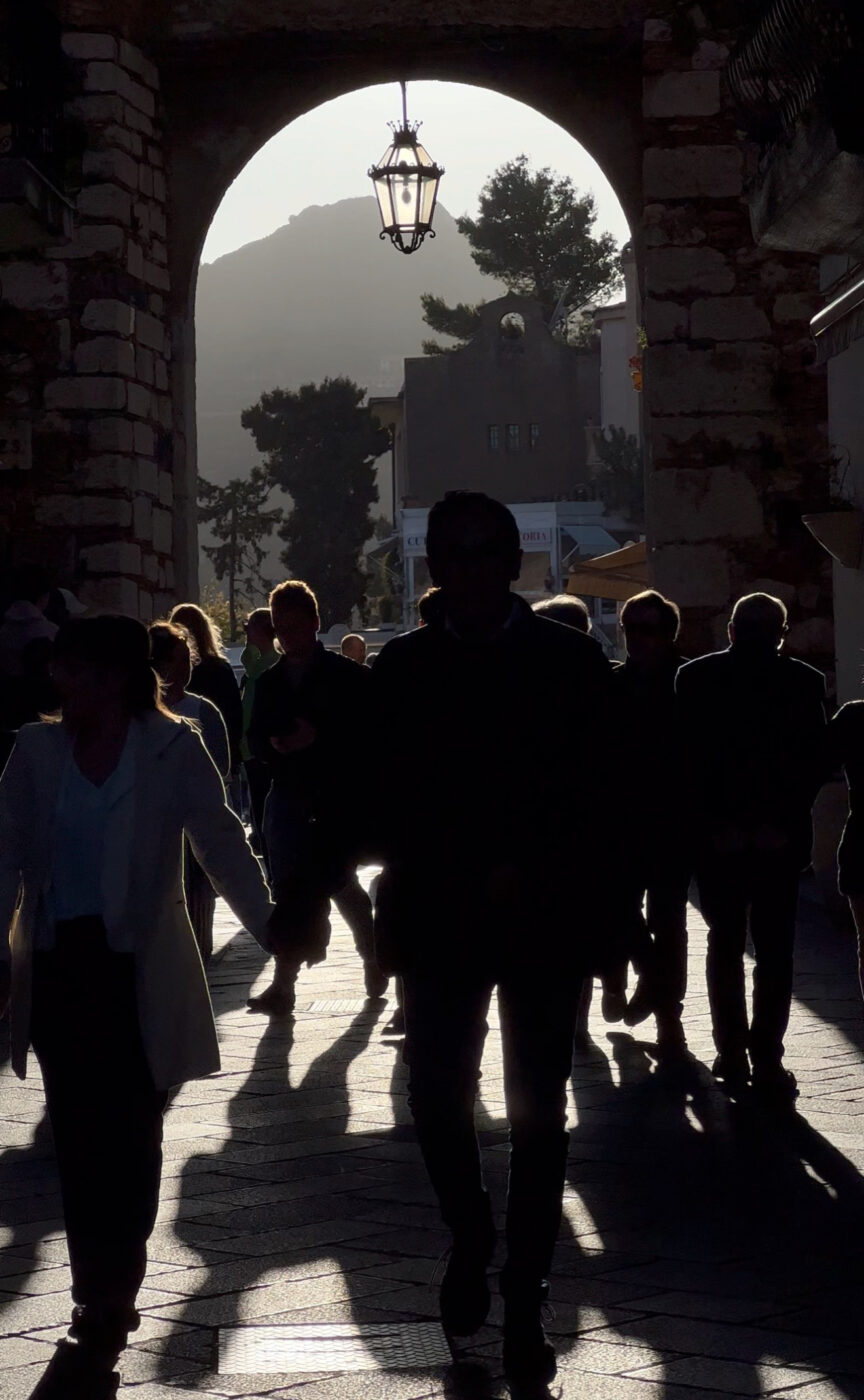 Sunset in Taormina - Silhouettes of people walking through a stone archway at sunset, with sunlight casting long shadows and a lantern hanging above. Trees and buildings are visible in the background.