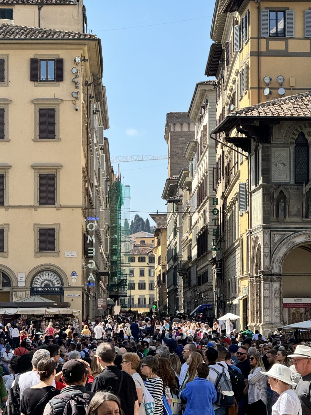 Piazza Duomo Florence - A large crowd fills a sunlit street lined with historic yellow buildings in Florence, Italy; the distant dome of the Cathedral of Santa Maria del Fiore is visible above rooftops.