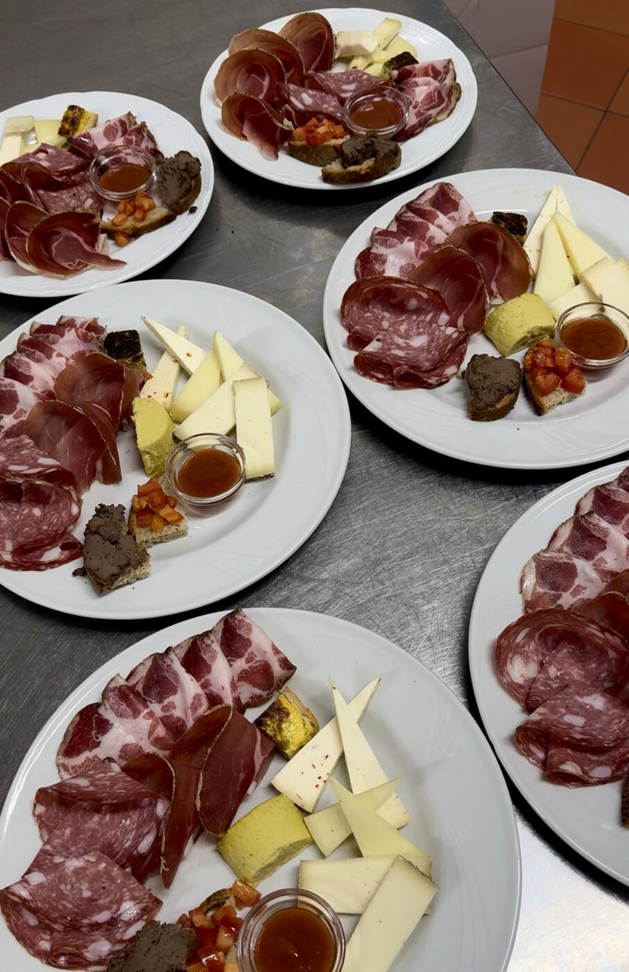 Five white plates on a metal surface - Each plate contains assorted cured meats, cheese wedges, pâté, small bread pieces, a tomato mixture, and a small cup of honey or sauce all arranged neatly.
