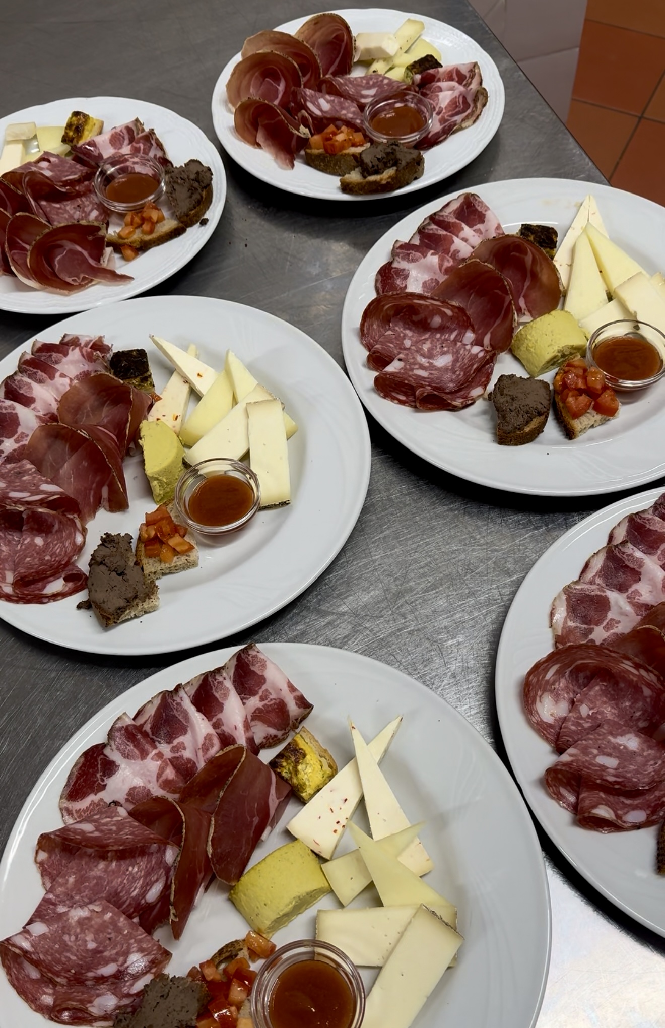 Five white plates on a metal surface - Each plate contains assorted cured meats, cheese wedges, pâté, small bread pieces, a tomato mixture, and a small cup of honey or sauce all arranged neatly.