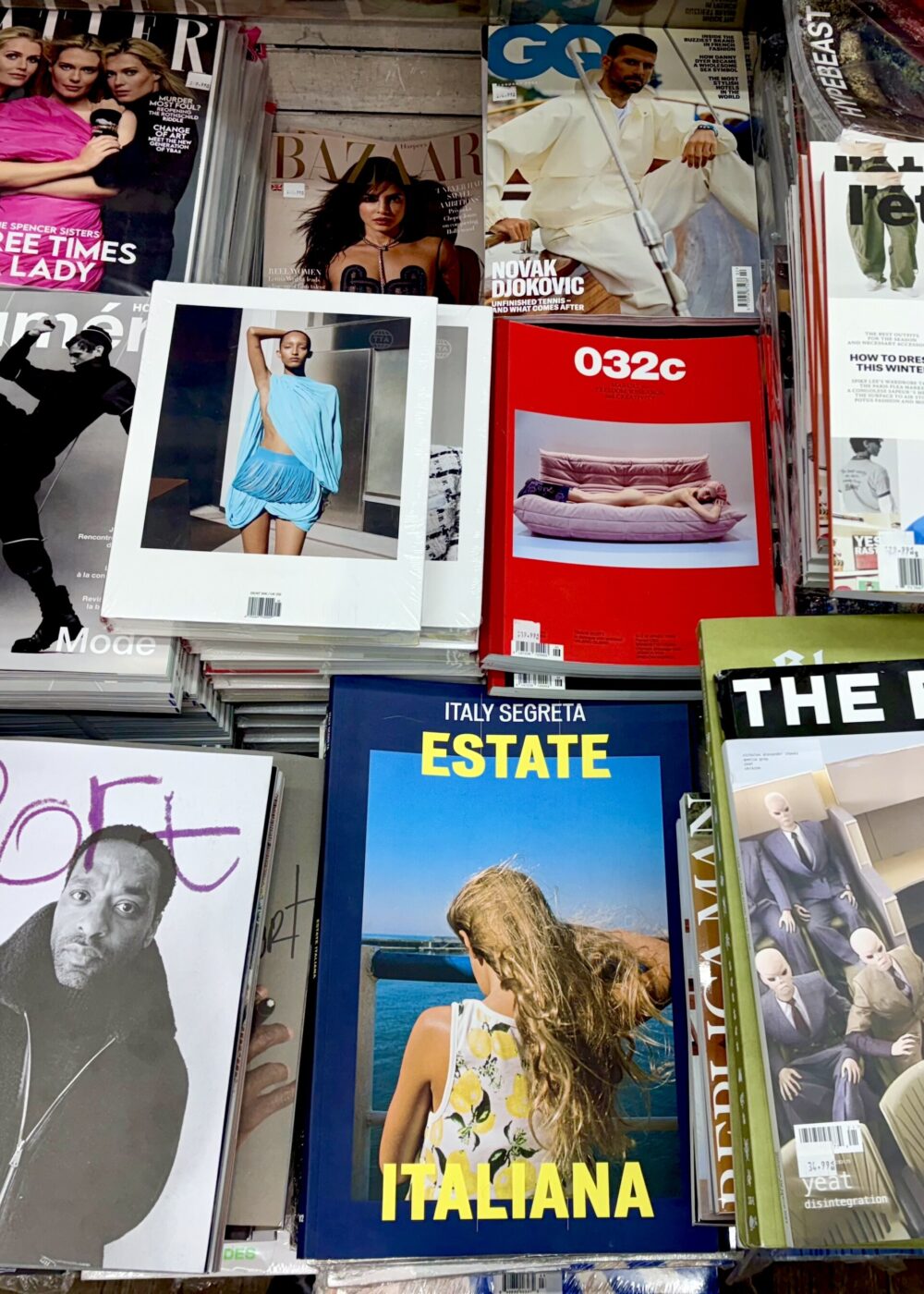 Magazine Display" - Un'esposizione di varie riviste su uno scaffale. Le copertine visibili presentano modelli, celebrità e testo in grassetto, con una varietà di colori e stili vivaci, tra cui temi di moda e stile di vita.
