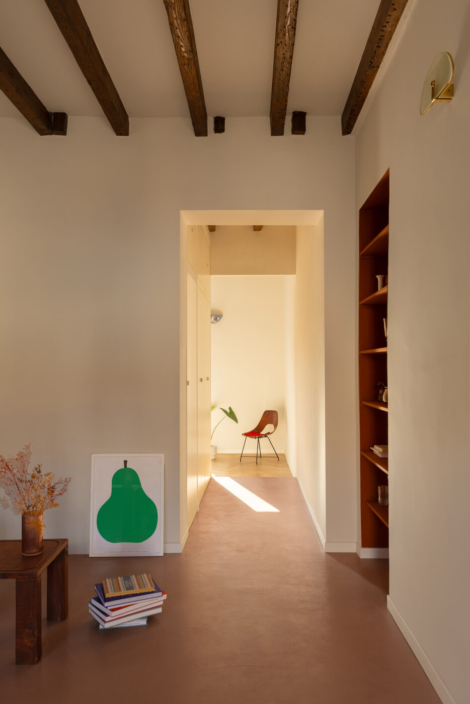 Camera minimalista con opere d'arte raffiguranti pere: una camera minimalista con travi in legno a vista sul soffitto, un corridoio illuminato dalla luce del sole che filtra dentro, una sedia in legno, un tavolino con un vaso, una pila di libri accanto e un'opera d'arte raffigurante pere verdi incorniciata appoggiata sul pavimento.